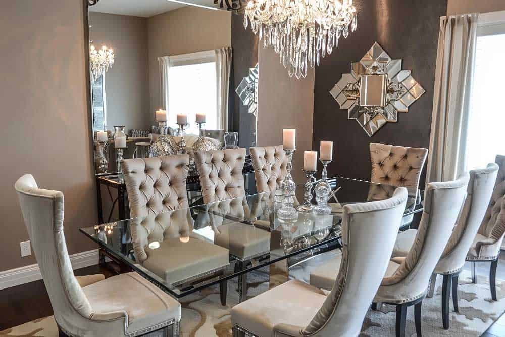 La imagen muestra un comedor elegante con mesa de cristal, sillas capitonadas en tonos claros y una lámpara tipo candelabro. Los espejos decorativos y la iluminación cálida crean un ambiente sofisticado, moderno y lleno de lujo.