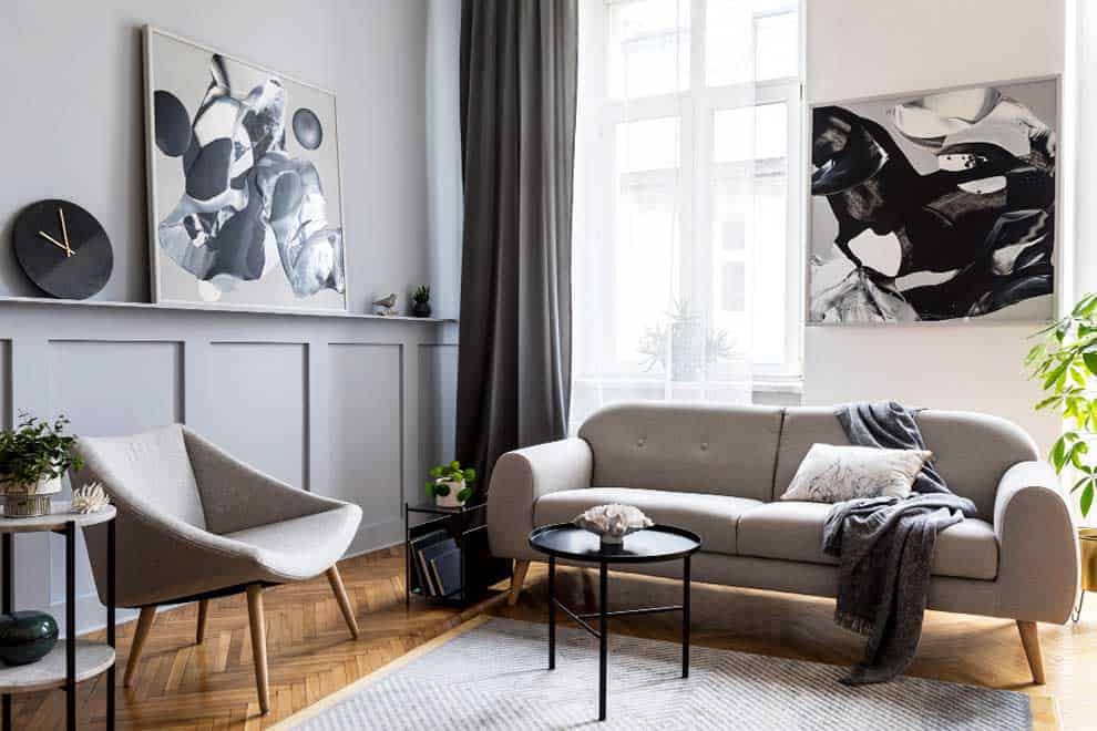 Sala contemporánea decorada en tonos grises y blancos. Destacan un sofá de líneas curvas, un sillón moderno, mesas de centro circulares con estructura metálica negra y cuadros abstractos que complementan un ambiente sofisticado.