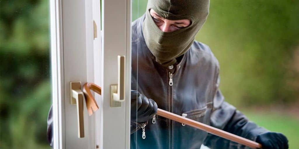 Se muestra a una persona con el rostro cubierto intentando forzar la apertura de una puerta con una herramienta metálica. La imagen representa un intento de robo a una vivienda y resalta la importancia de contar con cerraduras seguras.