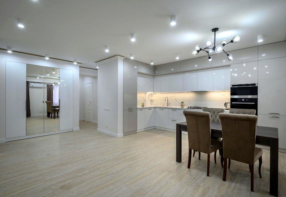 Se muestra una cocina integral moderna en color blanco, acompañada de un comedor de madera oscura con sillas tapizadas en tono beige. También se aprecia una iluminación empotrada en el techo y amplios espacios que aportan amplitud.