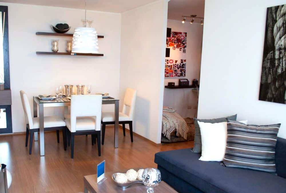 La imagen muestra un espacio integrado de sala y comedor con estilo moderno en tonos neutros. La distribución abierta conecta ambientes de forma funcional, mientras la decoración sencilla y la iluminación crean un entorno cálido.