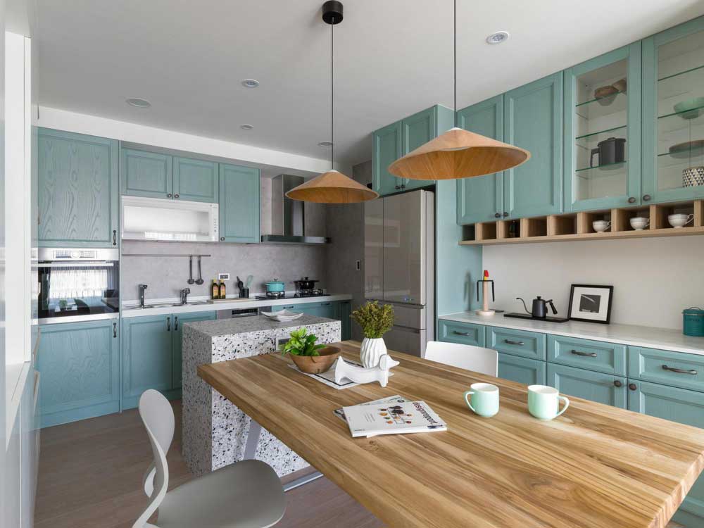 La imagen muestra una cocina moderna de estilo contemporáneo con gabinetes en color verde agua, una isla central con acabado tipo granito y un comedor de madera integrado.