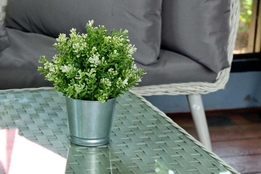La imagen muestra una mesa de centro con acabado tipo ratán y cubierta de cristal, decorada con una pequeña planta en maceta metálica. Al fondo se observa un sillón en tono gris que aporta un ambiente fresco, natural y acogedor.