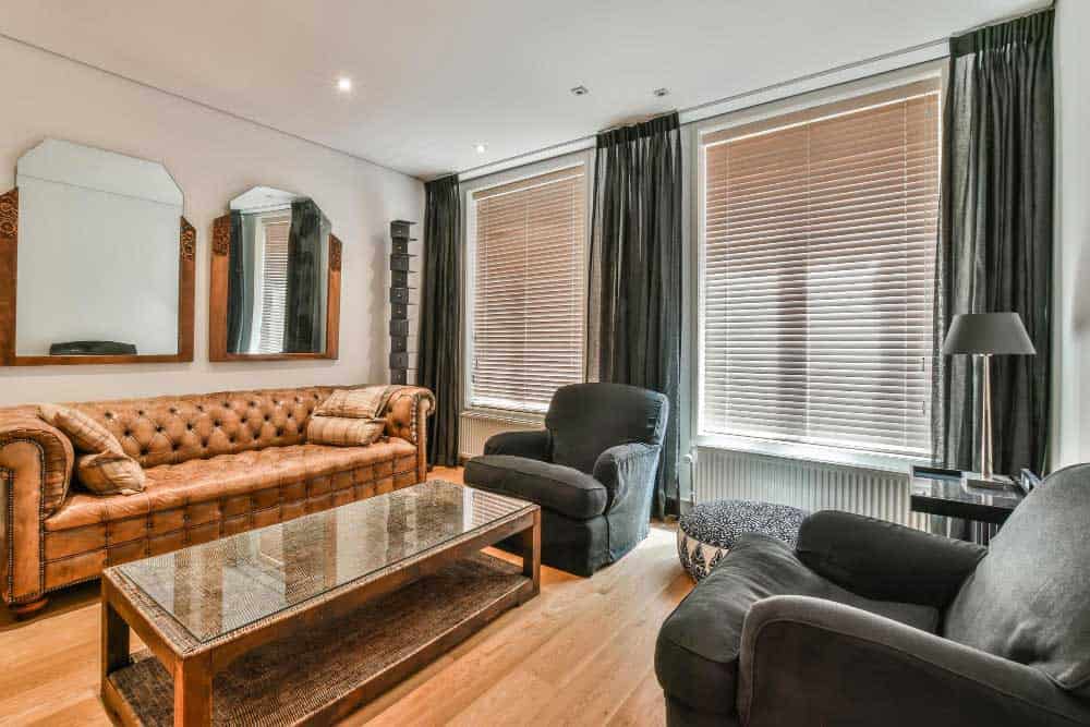La imagen muestra una sala de estar con estilo clásico y toques contemporáneos, destacando un sofá capitoné en tono café, sillones en gris y mesa de centro de madera. La iluminación natural y los espejos aportan elegancia, calidez y un ambiente acogedor.