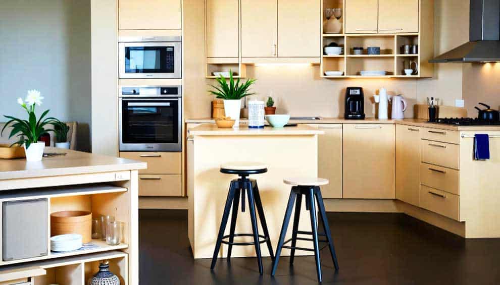 La imagen muestra una cocina integral moderna en tonos madera clara, equipada con una isla central y bancos altos. Su diseño aprovecha el espacio de manera funcional, combinando almacenamiento, superficies de trabajo y un ambiente acogedor.