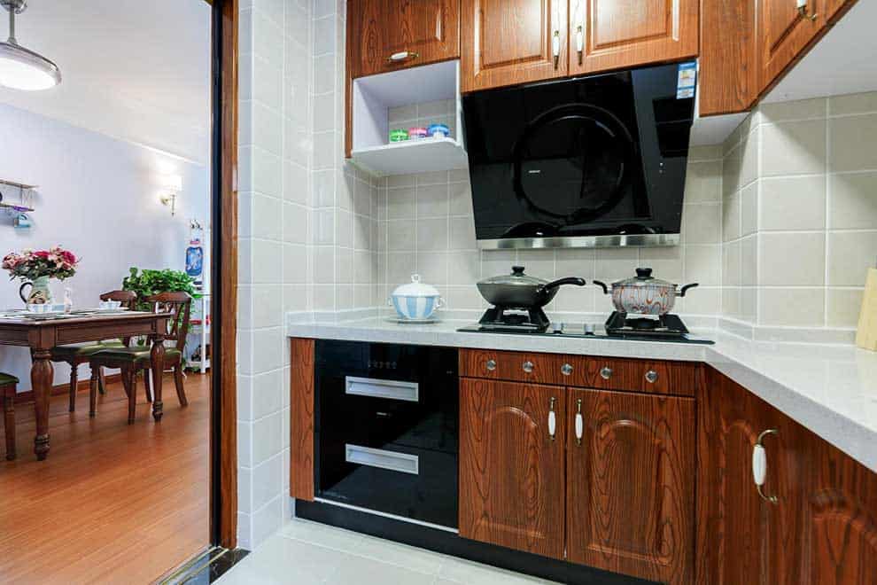 Cocina compacta con muebles de acabado tipo madera en tono oscuro y cubierta blanca. La campana negra, los electrodomésticos empotrados y la conexión directa con el comedor hacen que el espacio resulte práctico y perfectamente integrado al hogar.