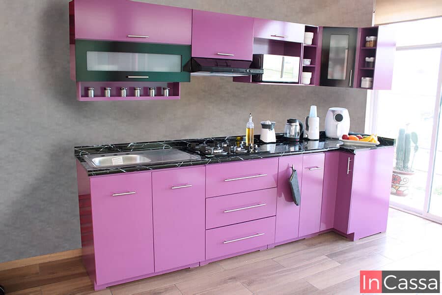 La imagen muestra una cocina moderna en acabado lila brillante con cubierta negra y múltiples compartimentos de almacenamiento. Su diseño aporta personalidad, color y funcionalidad a espacios contemporáneos.