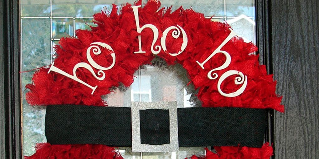 Se observa una corona navideña inspirada en el traje de Santa Claus, elaborada con malla decorativa en color rojo intenso. Destaca un cinturón negro con hebilla plateada al centro y la frase “ho ho ho” en la parte superior.