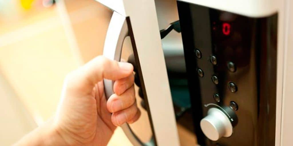 La imagen muestra una mano abriendo la puerta de un horno de microondas. Refleja el uso diario de este electrodoméstico para calentar o preparar alimentos rápidamente.