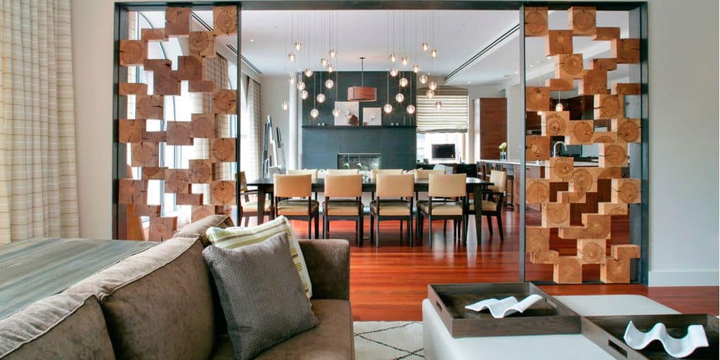 La imagen muestra un espacio moderno con sala y comedor integrados, separados por paneles decorativos de madera. Los tonos cálidos, la iluminación colgante y el diseño elegante crean un ambiente sofisticado, amplio y armonioso.