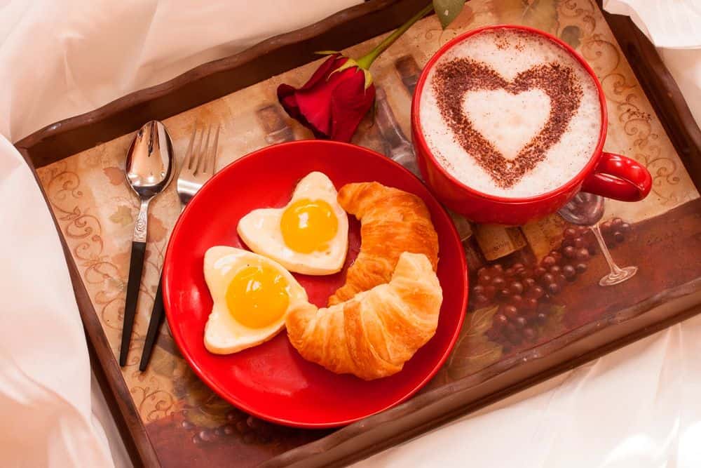 Se muestra un desayuno servido en la cama con un café decorado en forma de corazón, huevos con presentación creativa, un croissant y una rosa roja. La escena transmite un ambiente romántico y cálido, ideal para consentir.