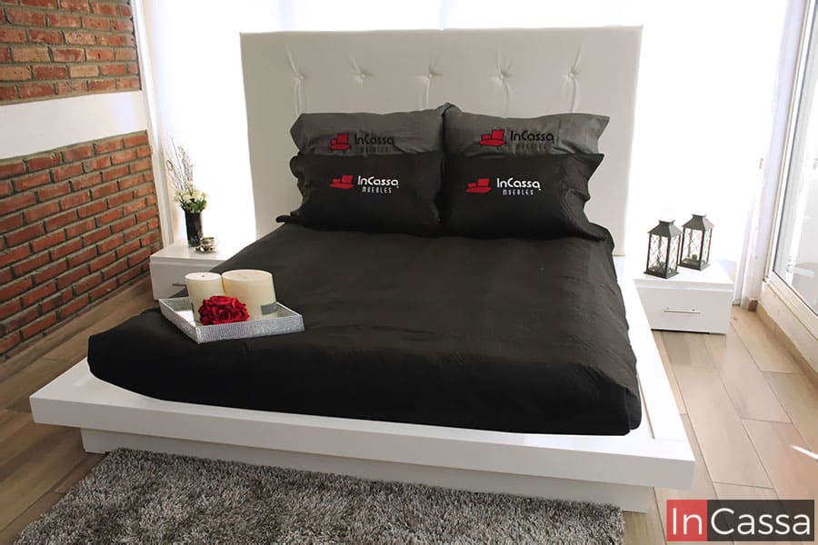 En la imagen se observa una recámara con cama de plataforma en acabado blanco, cabecera tapizada y burós laterales. La ropa de cama en color negro genera un elegante contraste con los muros de ladrillo aparente y la decoración minimalista.