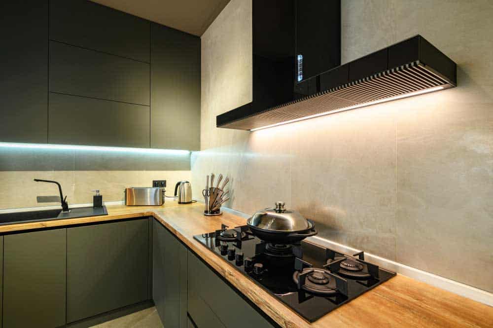La imagen muestra una cocina integral moderna en tonos oscuros con cubierta de madera natural y parrilla empotrada. Destacan la campana minimalista negra, iluminación LED indirecta y acabados mate que aportan un estilo elegante y sofisticado.