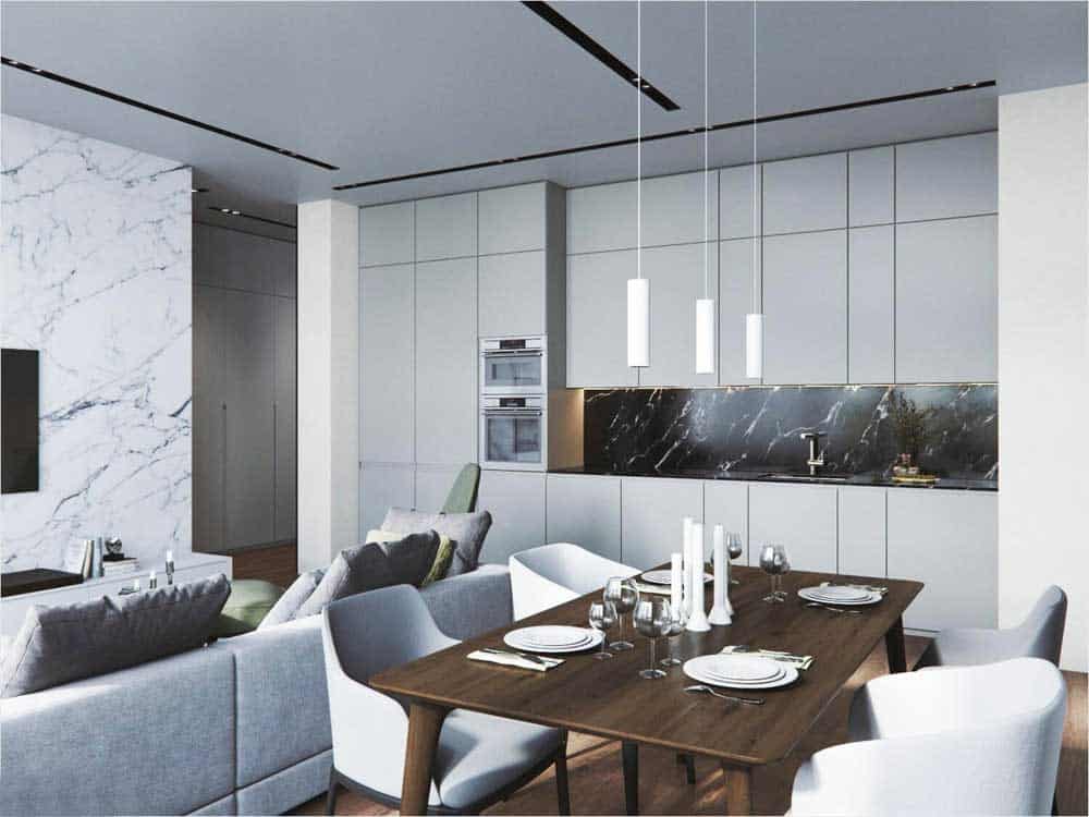 La imagen muestra una cocina integral abierta con comedor integrado y acabados minimalistas en tonos claros. Destacan los gabinetes de piso a techo, salpicadera tipo mármol oscuro y lámparas colgantes que aportan un estilo moderno y elegante.