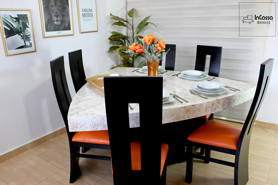 La imagen muestra un comedor moderno con mesa triangular de acabado claro tipo mármol, acompañado de sillas negras con asiento en tono naranja. La ambientación incluye vajilla elegante y centro floral, creando un espacio cálido y sofisticado.