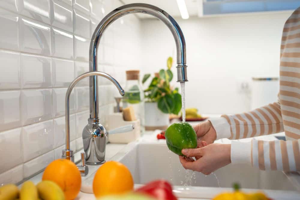 Se observa una cocina moderna donde una persona lava una fruta bajo un grifo de acero inoxidable. La iluminación natural y los detalles frescos crean un ambiente limpio, funcional y acogedor, ideal para el día a día.