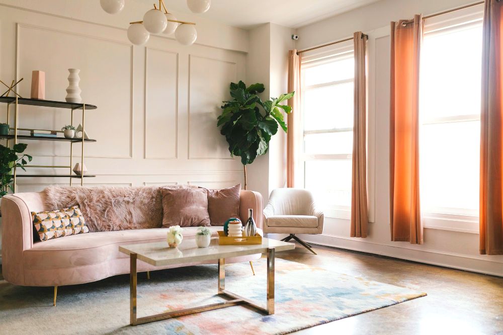 La imagen muestra una sala elegante con sofá en tono rosa, mesa de centro moderna y decoración en tonos cálidos. La luz natural y los detalles decorativos crean un ambiente acogedor, sofisticado y armonioso.