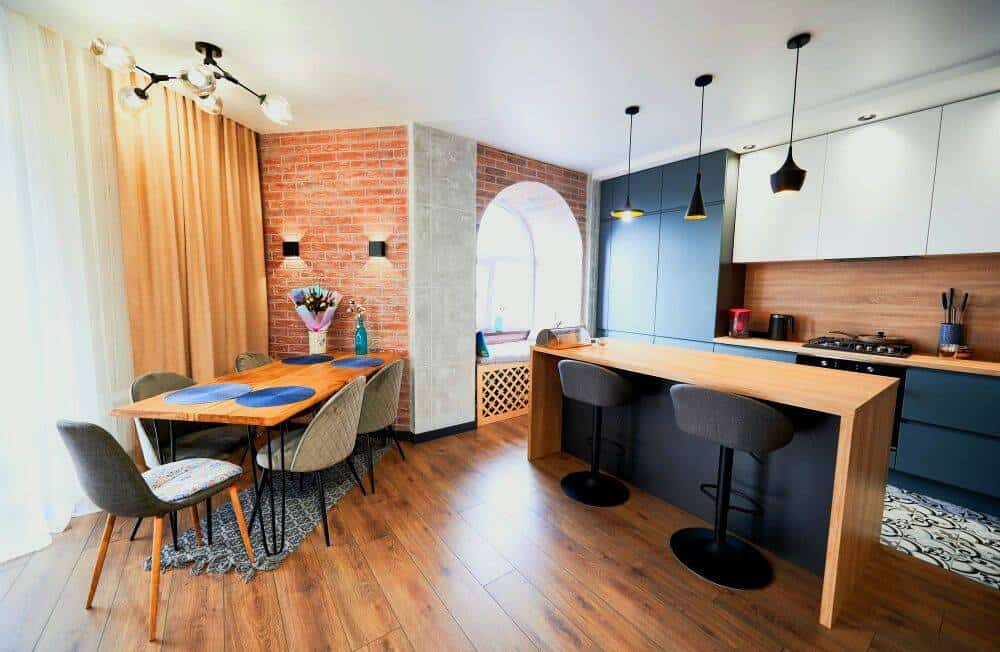 Espacio abierto que integra comedor y cocina, destacan una isla de cocina con barra desayunadora, un comedor de madera y una combinación de acabados en ladrillo, madera y tonos oscuros que aportan un estilo moderno e industrial.