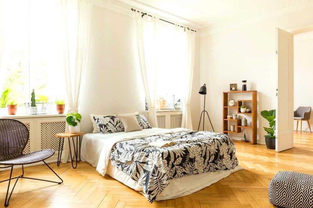 Habitación de estilo escandinavo con abundante luz natural, una cama vestida en tonos blanco y negro con estampado botánico, piso de madera y mobiliario minimalista. Las plantas, el estante decorativo y los textiles crean un ambiente cálido.