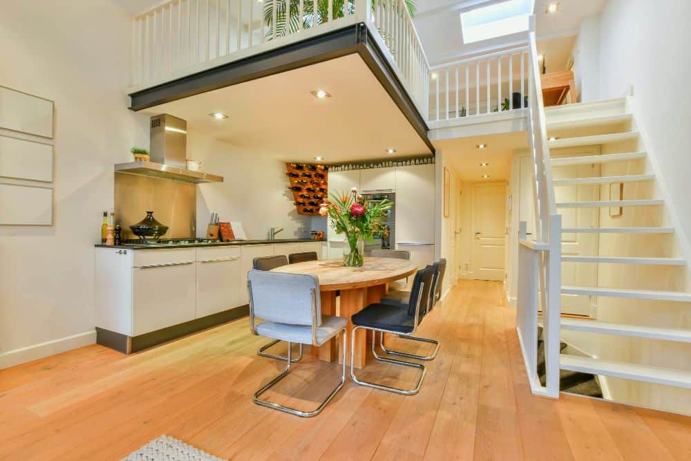 Espacio de concepto abierto que integra cocina, comedor y un nivel superior tipo mezzanine. Predominan los tonos claros, la madera natural y una iluminación cálida. La escalera blanca y los techos altos aportan amplitud y modernidad al ambiente.