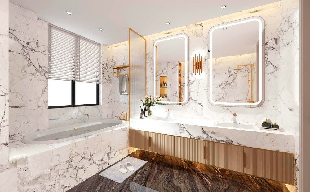Se muestra un baño de estilo lujoso revestido con superficies que imitan mármol blanco con vetas grises. Cuenta con una tina empotrada, doble espejo iluminado, amplio mueble de lavabo y detalles metálicos en color dorado.