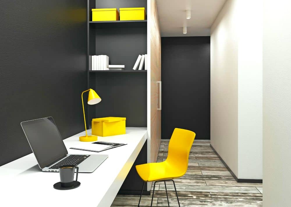Se observa un escritorio compacto instalado en un espacio reducido, aprovechando al máximo un pasillo o rincón estrecho. Destacan los contrastes en negro, blanco y amarillo, así como los estantes superiores que aportan almacenamiento.