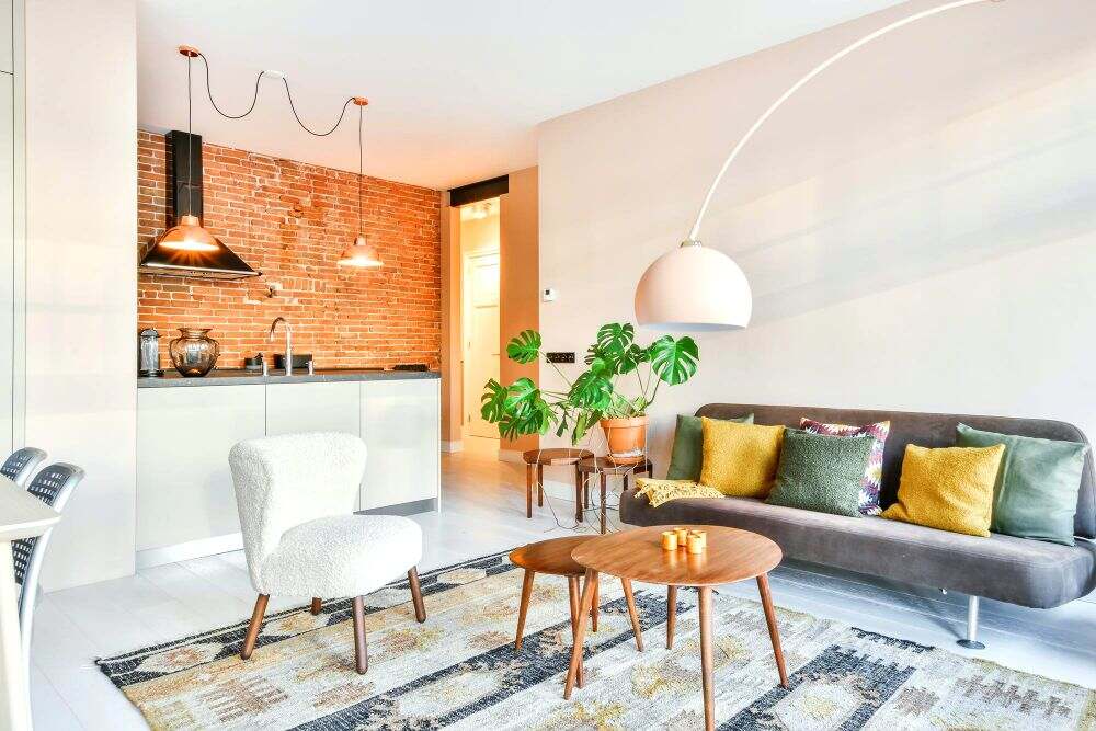 En la imagen se aprecia una sala moderna de concepto abierto que conecta con una cocina. Los muebles en tonos neutros, las mesas de centro de madera, las plantas decorativas y la iluminación cálida crean una atmósfera elegante y confortable.