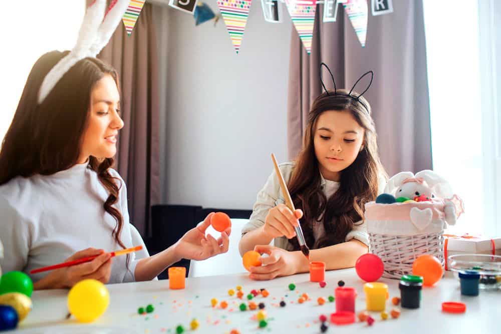 En la imagen se observa a una mujer y una niña decorando huevos de Pascua con pintura de colores. La mesa está llena de materiales para manualidades, creando un ambiente alegre, creativo y familiar.