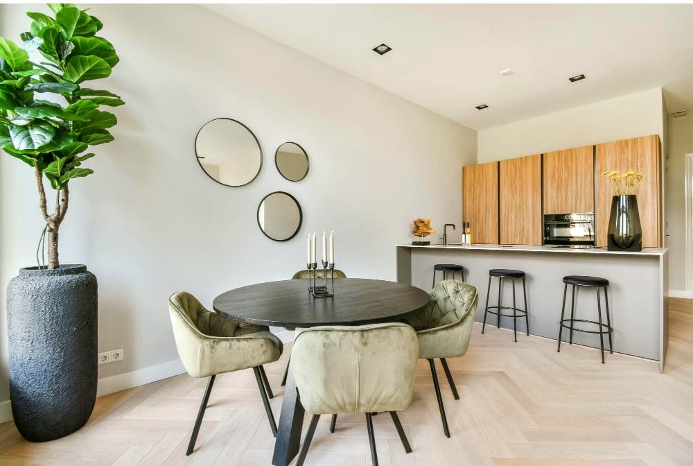 Comedor moderno con una mesa redonda en color negro y cuatro sillas tapizadas en tono verde olivo. Se aprecia una barra desayunadora con bancos altos, detalles en madera natural y una decoración minimalista con espejos circulares.