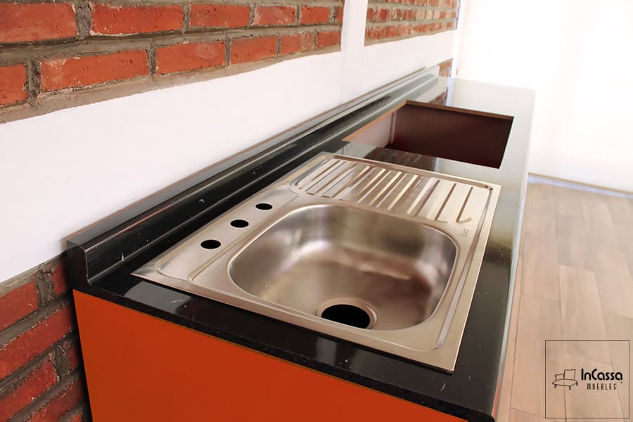 La foto se concentra en la cubierta de formica de la cocina, destacando principalmente la tarja de la cocina, la cual es de acero inoxidable y cuenta con escurridor del lado derecho. La imagen hace posible apreciar de mejor manera el acabado mármol negro de la cubierta, además de que es posible ver en la cubierta el hueco hecho para la instalación de una parrilla.