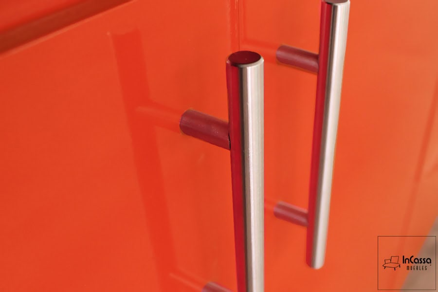 Está imagen se concentra en las jaladeras metálicas incorporadas en las puertas, cuyo diseño es en forma de "T" lo que da un aspecto mas moderno y estético a la cocina. Gracias a las jaladeras, es posible apreciar detalladamente el acabado brillante en la pintura naranja de la cocina.
