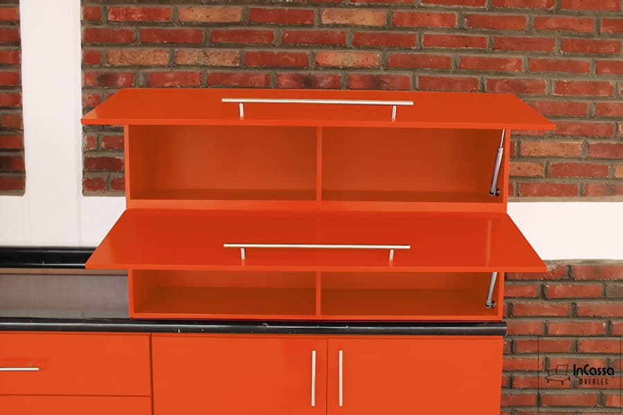 Foto de unos de los módulos superiores de la cocina, presentado con sus dos puertas abiertas dejando ver los cuatro compartimentos con los que cuenta. Todas las partes internas mantienen el mismo color naranja que se aprecia en las partes exteriores. También es posible ver los pistones neumáticos y las jaladeras integradas a cada puerta.