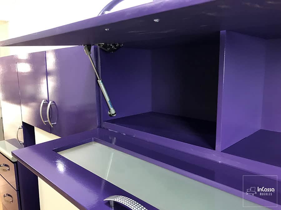 La imagen muestra los tres módulos superiores desde una toma lateral mostrando mayormente el módulo sobre tarja, el cuál destaca por sus pistones neumáticos integrados para una apertura y cierre vertical en sus puertas. Los compartimentos del módulo son completamente morados al igual que el resto de la cocina, la cercanía de la foto permite apreciar también el acabado brillante en la pintura de las partes exteriores de todos los módulos.