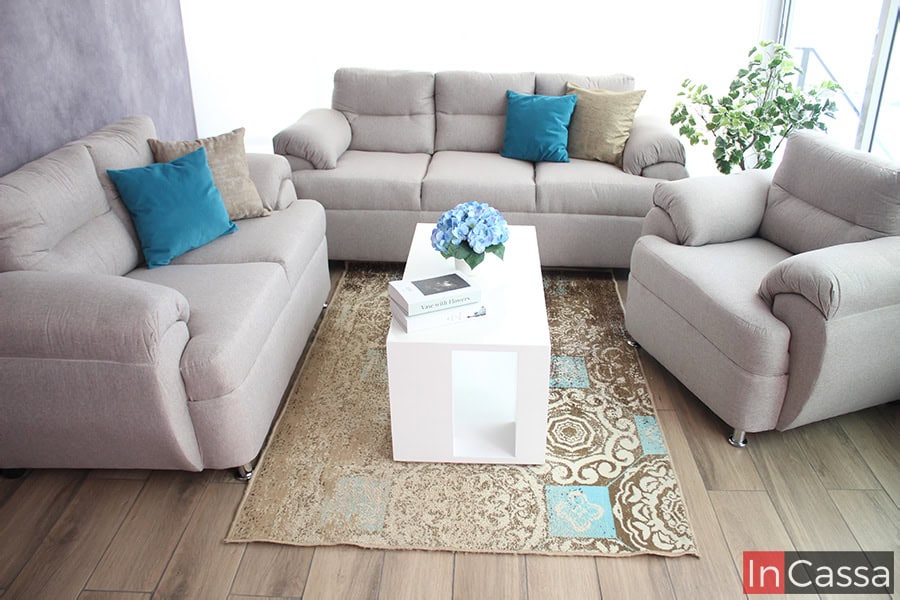 La imagen muestra la sala completa conformada por un sofá de tres plazas, un love seat y un sillón, todos tapizados en tela lino color greige y decorados con algunos cojines pequeños de tonos dorados y turquesa.