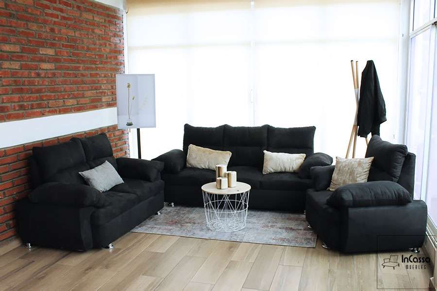 Una toma de la sala en tapiz suede negro decorada con algunos cojines decorativos de tonos hueso. Los tres módulos que conforman la sala se encuentran instalados en una estancia con pared de ladrillo y piso de madera.
