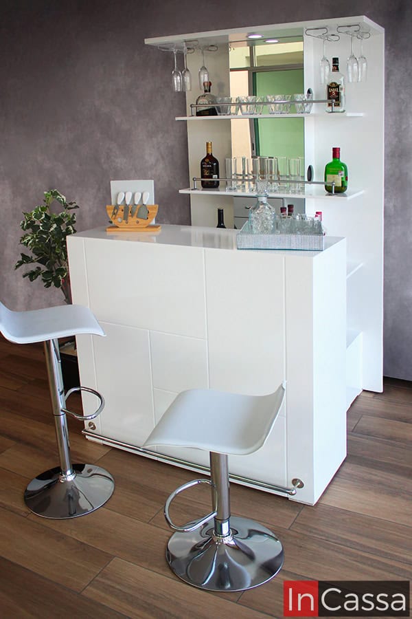 Un juego de cantina completo en color blanco. Este cuenta con un módulo trasero alto con espejo y repisas para bebidas y cristalería, además de una barra con patrón de formas geométricas y dos bancos modernos altos con asientos blancos. La cantina se encuentra en una estancia con pared gris y piso de madera.