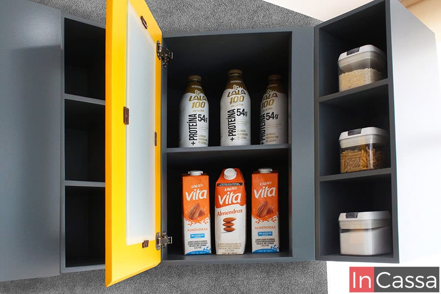 Se presenta un gabinete superior esquinero con puerta central en color amarillo cromo y marco de vidrio, acompañado de compartimentos abiertos en color gris medio a ambos lados. Ideal para almacenar alimentos de forma accesible y con un toque moderno y decorativo.
