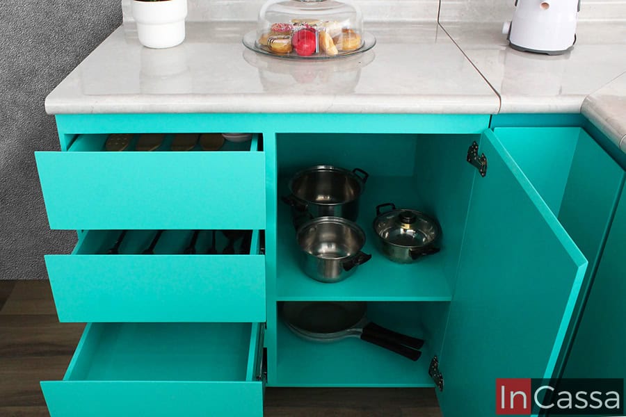 La imagen muestra un módulo inferior turquesa con tres cajones extensibles y un compartimento doble con puertas abiertas. Se observa la capacidad del mueble para organizar cubiertos y utensilios, así como para almacenar ollas y sartenes en su interior.