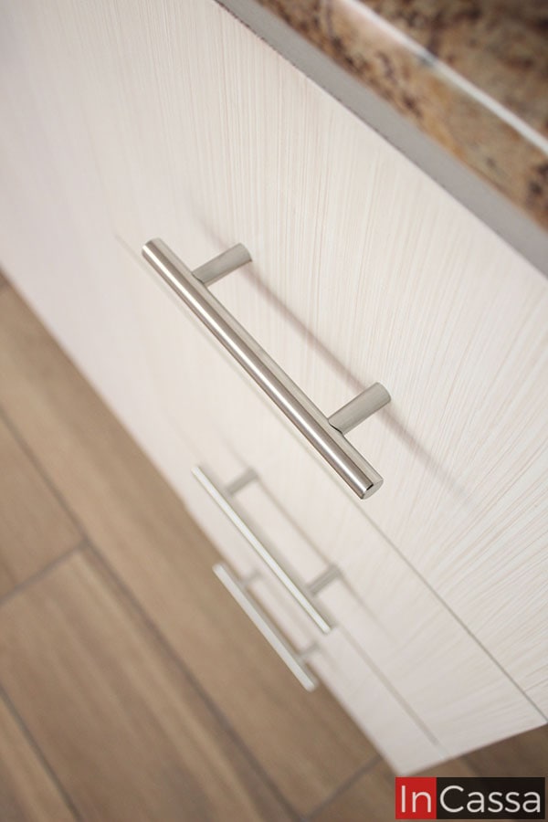 Una foto que da mayor prioridad a mostrar las jaladeras metálicas en estilo de barra T integradas a los cajones y puertas de cada módulo de la cocina. Además de que la cercanía de la toma, también permite apreciar el acabado laqueado de la pintura color paja presente en toda la cocina.