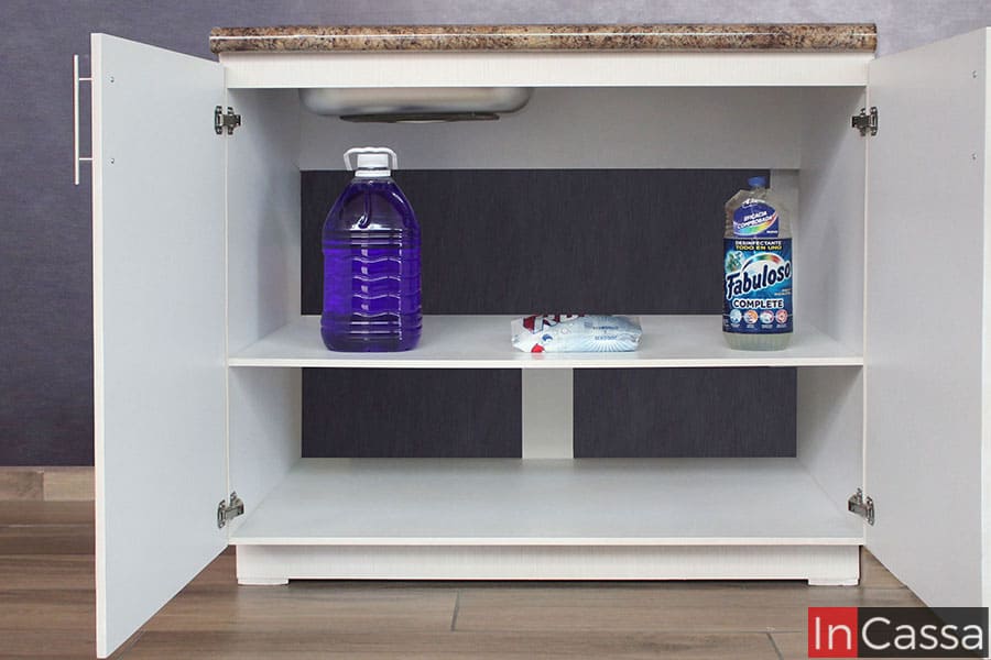 La foto muestra el mueble de tarja de la cocina con sus puertas abiertas, dejando a la vista dos compartimentos bastante amplios divididos por un entrepaño horizontal. El interior del módulo es blanco en su totalidad y mantiene un gran espacio descubierto para la instalación de plomería para la tarja.