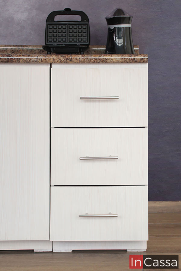 Foto de los tres cajones laterales del módulo inferior de la cocina; estos son del mismo tamaño y mantienen el acabado paja que puede verse en el resto de la cocina. Cada uno de los cajones tiene una jaladera de barra T para su apertura y cierre, además la toma permite apreciar parte de la cubierta de formaica de estilo cuarzo café.