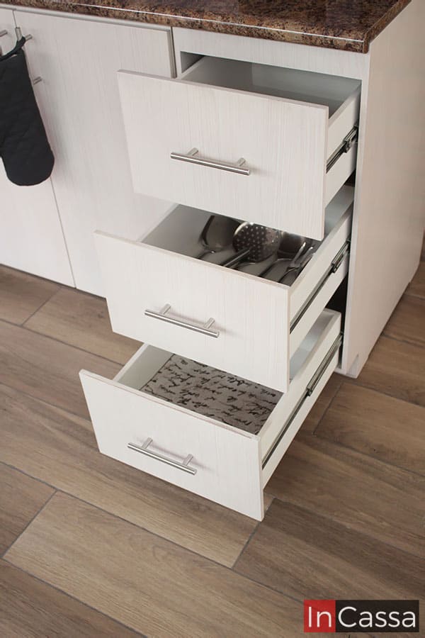 La imagen presenta los tres cajones laterales de la cocina abiertos, mostrando la totalidad de su estructura en la que resaltan sus rieles metálicos y sus jaladeras de barra T, mismos que conforman el sistema de apertura y cierre de cada cajón. Los tres compartimentos presentan un acabado paja en sus exteriores mientras que sus interiores son de color blanco.