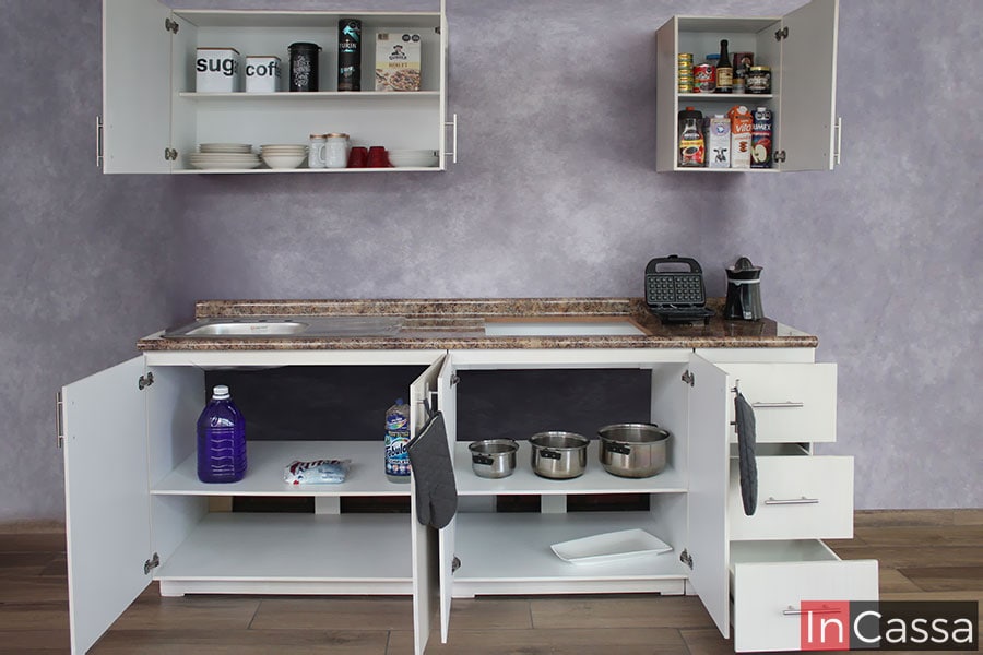 La cocina se muestra con todos sus compartimentos abiertos dejando ver los interiores de cada módulo, los cuales son de color blanco. También es visible el hueco para parrilla en la cubierta de formaica de estilo cuarzo café.