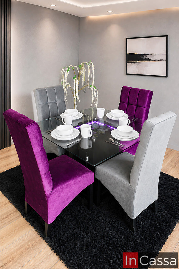 Se observa el Comedor Modelo AUSTIN para 4 personas con pedestal en color chocolate y cubierta de cristal, acompañado de sillas tapizadas en suede gris y morado con elegante acabado capitoneado. Su diseño moderno con iluminación integrada crea un ambiente sofisticado, ideal para realzar cualquier comedor y disfrutar momentos inolvidables con familia y amigos.