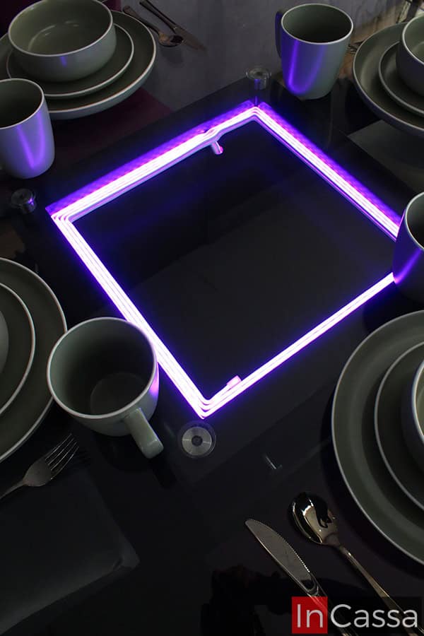Una foto del arenero de la mesa tomada en un ambiente con poca luz, a fin de resaltar la iluminación LED integrada al arenero de la mesa, la cuál es mostrada en color purpura, resaltando el juego de vajillas de porcelana blanca utilizado para la decoración de la mesa.