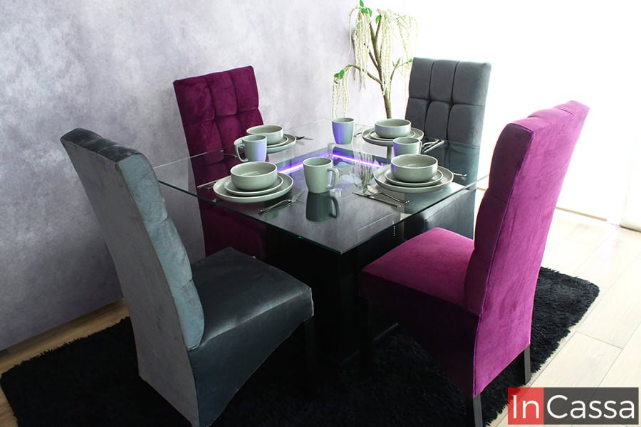 Una toma completa del comedor presentado en una estancia con pared gris, ventana de pantalla completa y piso de madera, en la que se resalta su mesa de cristal con iluminación LED y sus sillas tapizadas en suede gris y morado con acabado capitoneado.