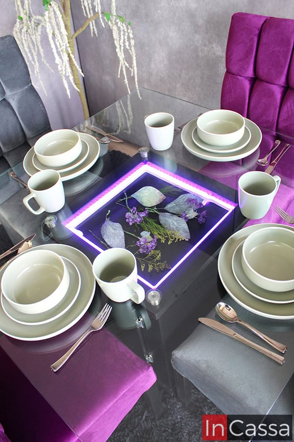 Una toma superior de la mesa del comedor, destacando su cubierta de cristal y su arenero adornado con flores y plantas decorativas, además de su iluminación LED mostrada en color morado. La mesa se encuentra decorada por un juego de vajillas de porcelana blanca y algunos cubiertos plateados.