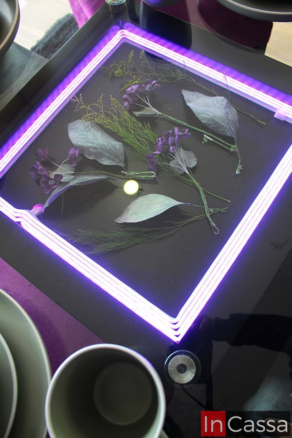 La imagen destaca el arenero con iluminación LED de la mesa, la cuál es mostrada encendida en color purpura. El arenero es presentado decorado con algunas flores y plantas decorativas, visibles gracias a la cubierta de cristal de la mesa.