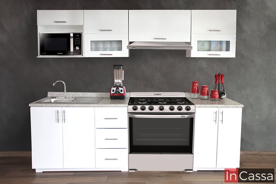 Cocina integral blanca para estufa de piso, la cuál consta de dos módulos inferiores con cubierta de formaica y tres módulos superiores entre los que resalta una alacena superior con espacio para microondas. La cocina se encuentra instalada en una estancia con piso de madera y pared gris.