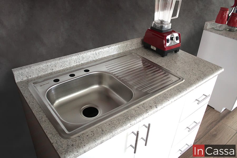 La toma se concentra en la tarja de acero inoxidable la cuál cuenta con tina y escurridor, y se encuentra integrada a la cubierta de formica estilo sal y pimienta del módulo de tarja de la cocina blanca.